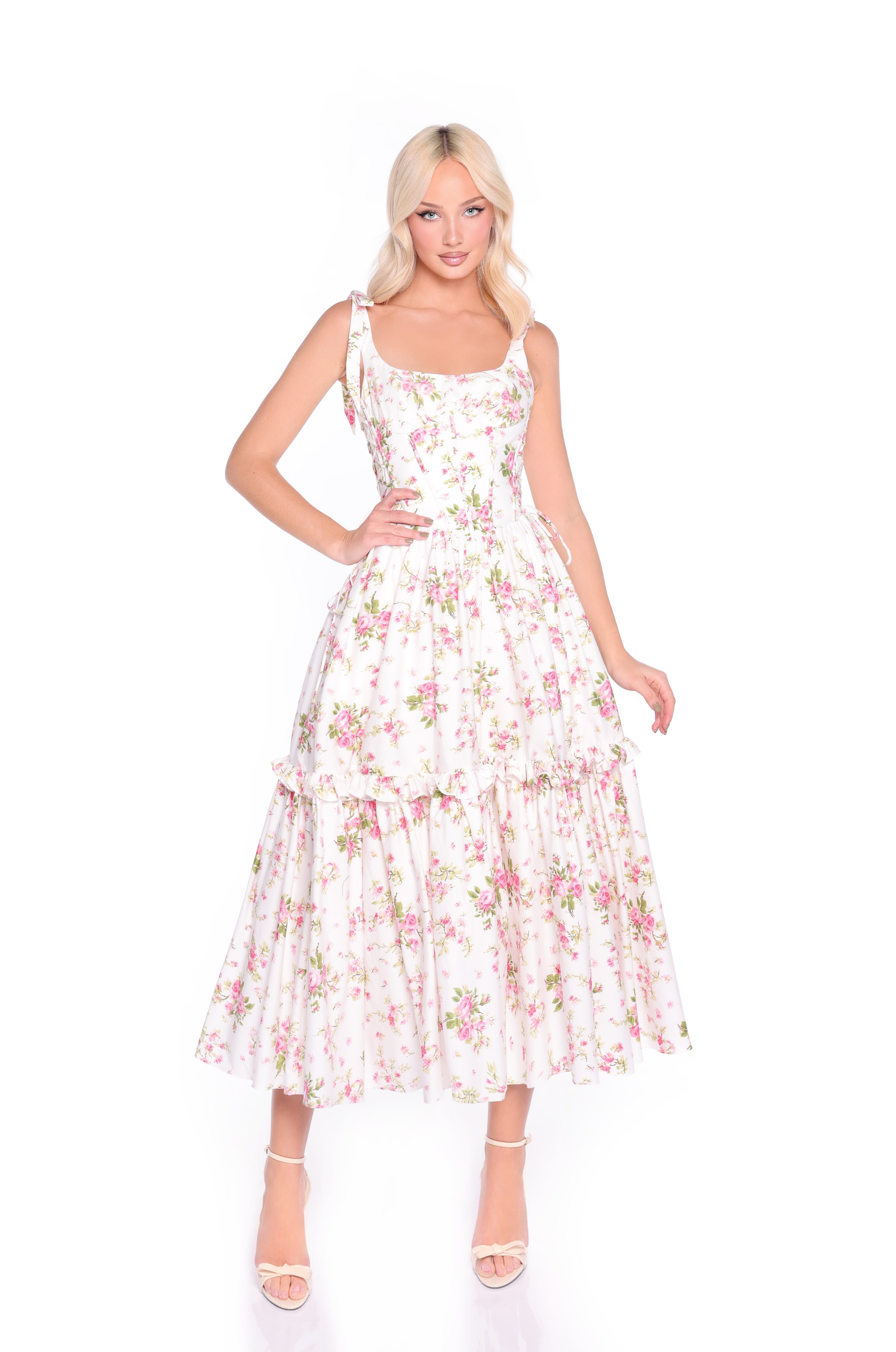 A woman poses in the Ivory Floral Casual Midi Corset Dress: Rosalie, a sleeveless fit-and-flare dress with pink floral patterns and ruffled tiers, paired with open-toe heels against a white background.